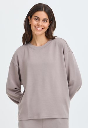 Jeune femme aux longs cheveux foncés sourit, portant un sweat-shirt ample violet clair et un pantalon assorti sur un fond blanc uni.