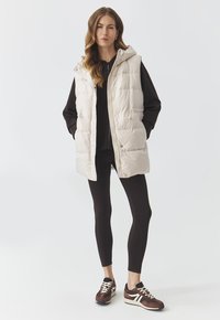 Padded beige vest with a hood, front zip, and snap buttons. Worn over a dark top and paired with black leggings and brown sneakers.