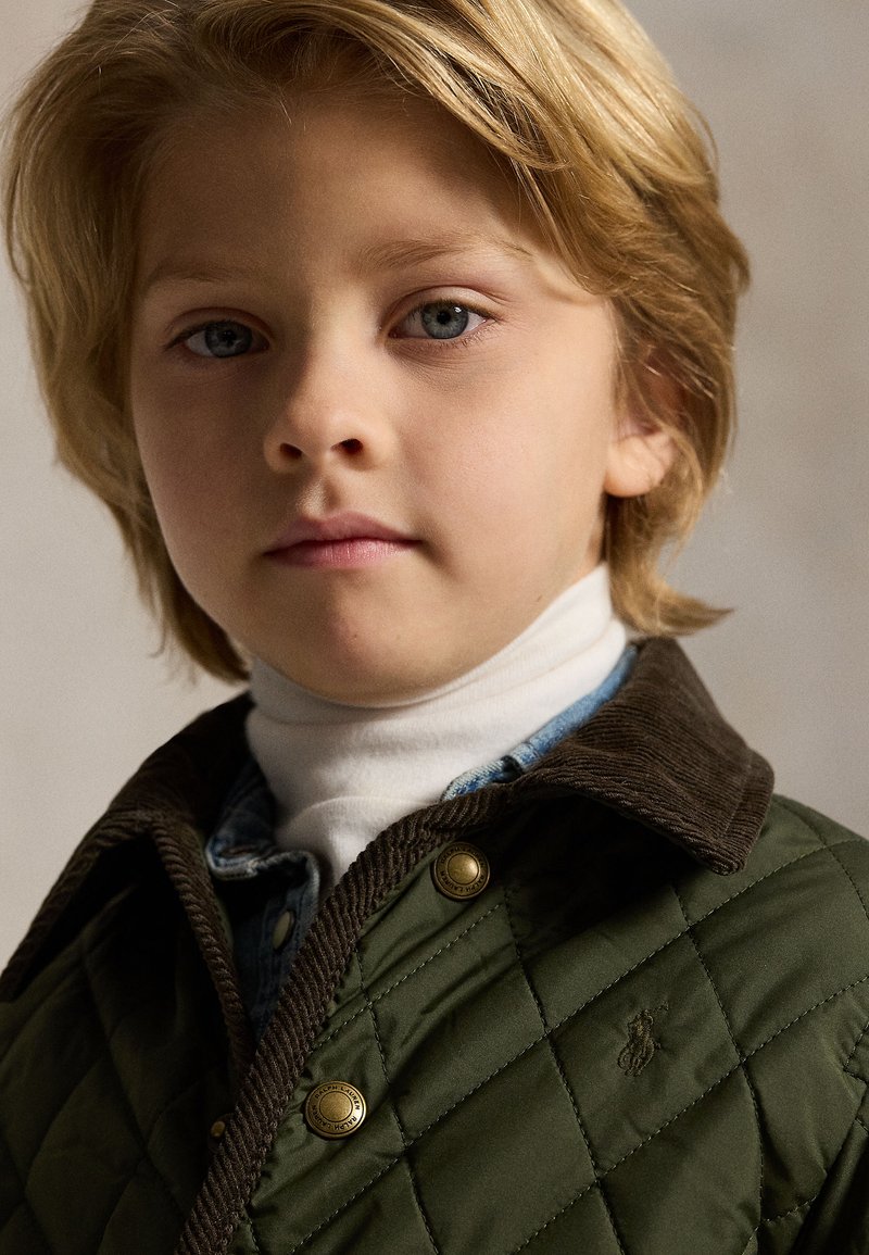 Niño pequeño con cabello rubio que lleva una chaqueta verde acolchada sobre un jersey de cuello alto blanco y una camisa vaquera, mirando hacia adelante con expresión neutral.