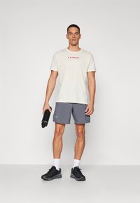 Ljusbeige t-shirt med röd "TRAIL"-logotyp, kombinerad med grå sportshorts. Håller en svart vattenflaska, bär svarta sneakers med vita strumpor.
