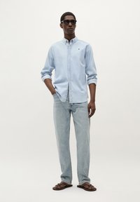 Lichtblauw button-up shirt, relaxte pasvorm. Gecombineerd met lichte spijkerbroeken en bruine flip-flops. Model draagt ook zwarte zonnebril.