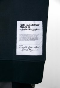 Tessuto nero con un'etichetta bianca prominente che presenta testo e firme. L'etichetta descrive un capo di moda collaborativo in edizione limitata.