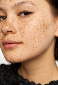 Närbild av ett ansikte med ljus hy som har många bruna fräknar, mjuka läppar och subtil eyeliner. Bakgrunden är mörk och texturerad.