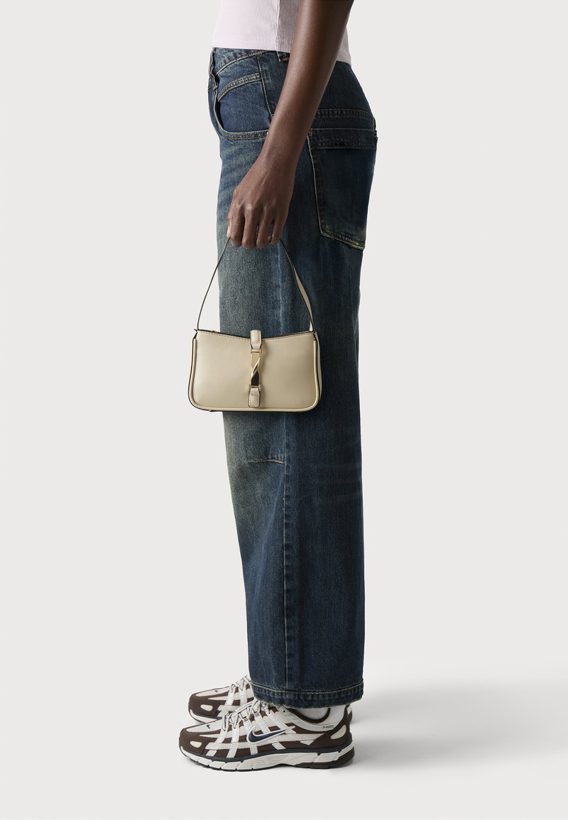 Beige nahkalaukku suorakulmaisen muodon kanssa, jossa on yläkahva ja keskellä oleva hihnadesign, pidetään henkilön käsissä, jolla on päällään tumman denim-kankaiset farkut ja urheilukengät.