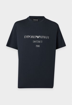 Νέος άνδρας που φοράει μαύρο t-shirt Emporio Armani, σκούρο παντελόνι και άσπρα αθλητικά παπούτσια στέκεται μπροστά σε μια απλή λευκή επιφάνεια.