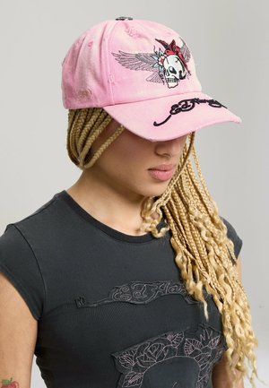 Osoba z długimi blond warkoczami, nosząca zniszczoną różową czapkę z projektem z skrzydlatą czaszką oraz czarną koszulkę Ed Hardy.