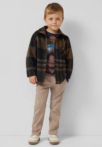 Koszula flanelowa w czarno-brązową kratę, zestawiona z granatowym t-shirtem z nadrukiem, beżowymi spodniami i białymi trampkami.