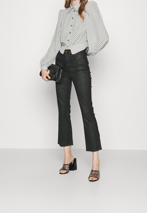 Kvinde iført en hvid bluse med mønster og pufærmer, sorte cropped flare bukser, sorte sandaler med hæl og holder en sort clutchtaske.