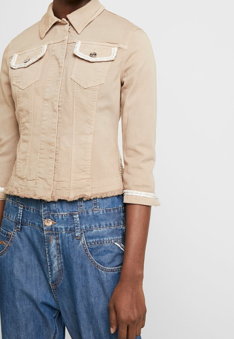 Beige denimjas met een kraag, driekwart mouwen en twee voorzakken, met contrasterende witte stiksels en een rafelige zoom.