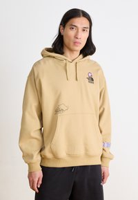 Felpa beige con cappuccio regolabile, tasca frontale e stampe grafiche, tra cui un'immagine di campeggio e testo colorato sulla manica.