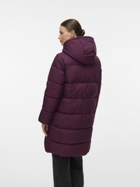 Sieviete, ģērbusies garā, pildītā purpursarkanā mētelī ar kapuci, stāv trīs ceturtdaļu atpakaļ skatā pret baltu fona.