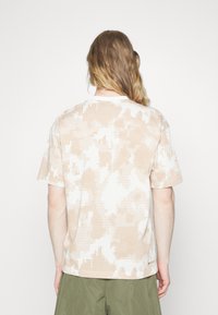 Beige och vit mönstrad kortärmad skjorta med pixelmönster, rund halsringning och avslappnad passform, tillverkad av lätt tyg.