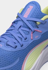 Κοντινό πλάνο μπλε αθλητικού παπουτσιού Puma με ροζ και κίτρινες λεπτομέρειες, με το κείμενο "Fit+" και χοντρές, υφασμάτινες κορδέλες.