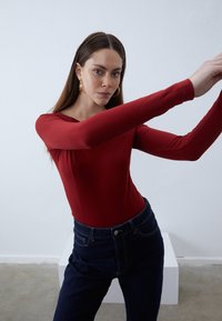 Long-sleeve red bodysuit made of smooth fabric, featuring a rounded neckline and fitted silhouette, paired with dark blue high-waisted jeans.