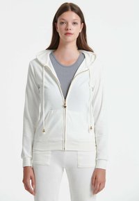 Bijela zip-up hoodie sa kapuljačom na vezanje, zlatnim detaljima na rajsferšlusu i prednjim džepovima. Nosena preko sive rebraste majice bez rukava. Mekana tkanina.