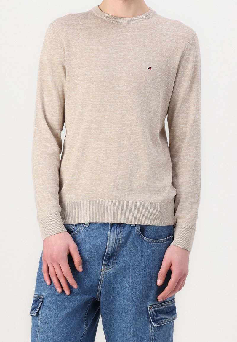 Pull beige à manches longues col rond avec petit logo sur la poitrine, porté avec un pantalon cargo en jean bleu.