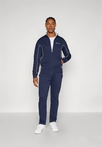 Mornarsko modra trenirka z belimi poudarki, ki vključuje jakno z zadrgo in hlače z elastičnim pasom. Izdelana iz mehke tkanine, dopolnjena z logotipom.