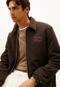 Młody mężczyzna ubrany w brązową bluzę zapinaną na zamek Tommy Hilfiger, na beżowym swetrze i białych spodniach, patrzy w lewo.