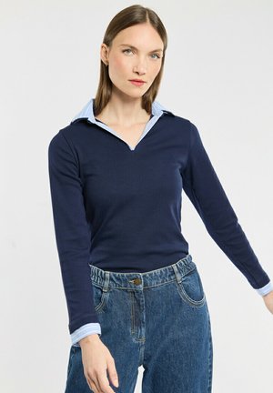Navy blauwe longsleeve top met een lichtblauwe kraaglaag, voorzien van een V-hals ontwerp en een gladde, gestructureerde stof. Gecombineerd met hoogproductie spijkerbroek.