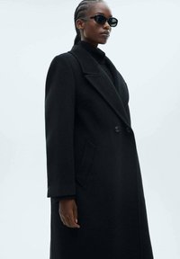 Cappotto doppiopetto nero in lana con colletto alto, dotato di tasche laterali e silhouette strutturata. Accompagnato da occhiali da sole scuri.