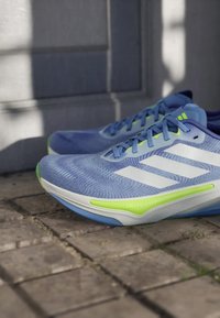 Sinivalkoiset lenkkitossut vihrein yksityiskohdin ulkoseinäpäällysteen päällä pehmeässä auringonvalossa.