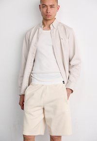 Light beige button-up shirt over a white tank top, paired with cream-colored shorts featuring side pockets and an Adidas logo.