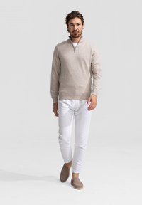 Beige ritsweater met kraag, witte slim-fit broek, bruine instappers. Model staat op een effen achtergrond en toont het eenvoudige ontwerp van de outfit.