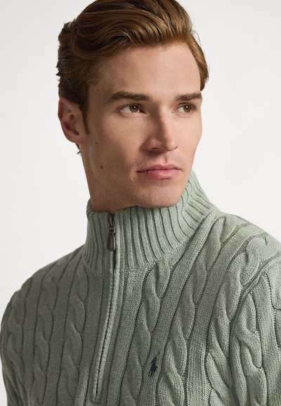 Knitted sweater in soft green with a high ribbed collar and zip closure. Features textured cable patterns and a small logo accent.