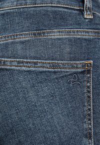 Jeans i denim med mörkblå färg. Egenskaper inkluderar en bakficka med orange sömnad och en liten broderad logotyp nära fickan.