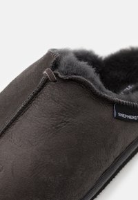 Donkerbruine suède pantoffel met zachte, fluffy zwarte voering en een "Shepherd" stoffen label aan de zijkant, getoond in close-up.