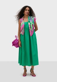 Grön bomullsmaxiklänning med rynkad midja, tillsammans med en färgglad blommig scarf och en rosa handväska. Klacksandaler med turkosa accenter.