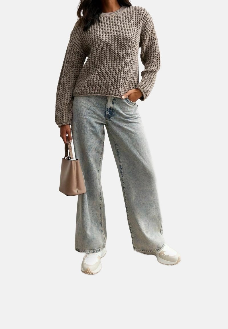 Gruby beżowy sweter, jasnoniebieskie szerokie dżinsy i białe sneakersy. Nosi małą beżową torebkę. Miękka tekstura i casualowy styl.