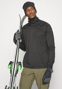 Camiseta térmica negra con media cremallera, guantes grises, esquís con detalles en verde y pantalones cargo de color oliva. El material parece ligero; diseñado para actividades invernales.