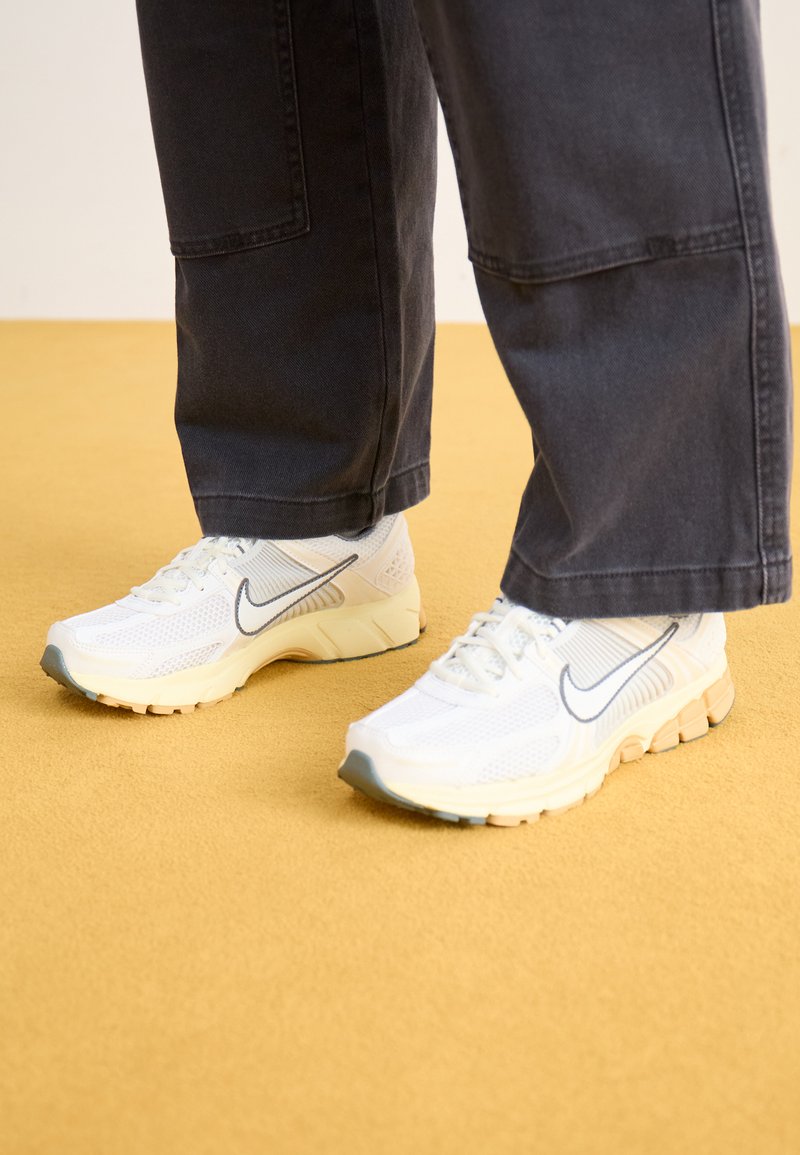 Witte sportschoenen van mesh en synthetische materialen, met een grijs logo, een gestructureerde zool en beige accenten, gedragen met donkere broeken.
