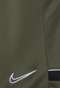 Gros plan sur un tissu vert olive avec le logo Nike swoosh blanc et une partie d’un détail noir et blanc.