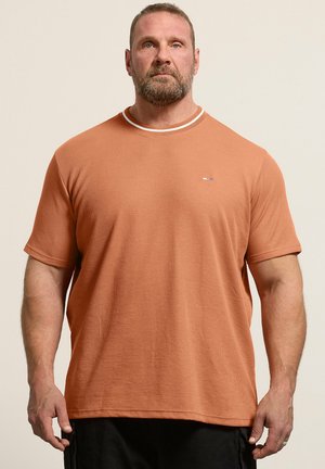 Brodaty mężczyzna w rudobrązowej koszuli z krótkim rękawem o subtelnej prążkowanej fakturze, stojący na jednolitym beżowym tle.