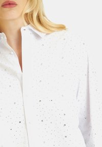 Camicia bianca con bottoni, decorata con embellishments in cristallo sparsi, collarino classico e una texture morbida.