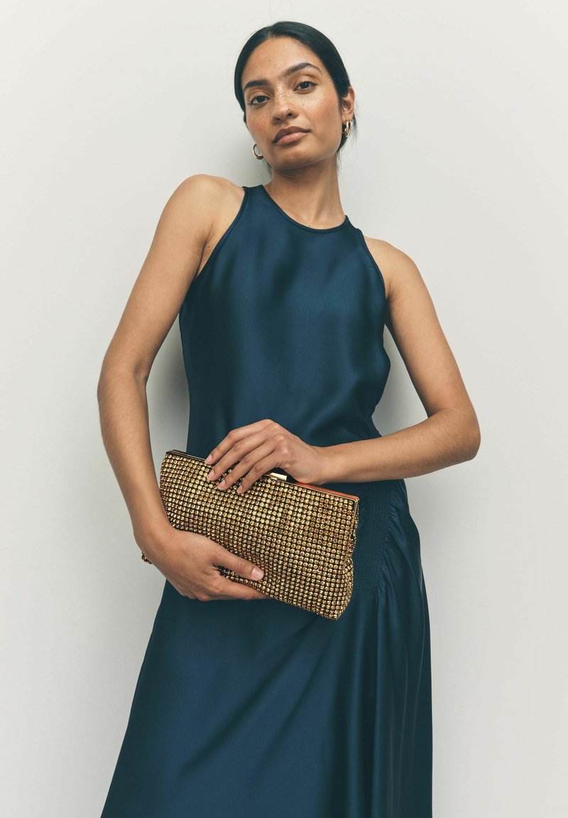 Mørkeblå satin uden ærmer kjole med en tætsiddende silhuet, parret med en guldfarvet clutch-taske med nitter, der har en topzip-lukning og tekstureret overflade.