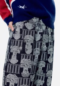 Pleteni plavi pulover s crvenim akcentima, uparen s sivim hlačama koje imaju ponavljajući uzorak klasičnih bisti i stupova. Teksturirani materijal.