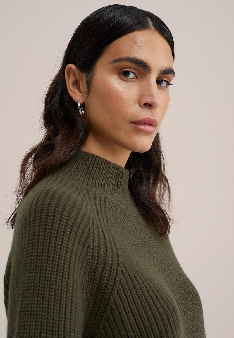 Oliwkowy sweter dzianinowy z wysokim kołnierzem, prążkowaną fakturą i dopasowanym krojem. Srebrne kolczyki w kształcie kół podkreślają stylizację.