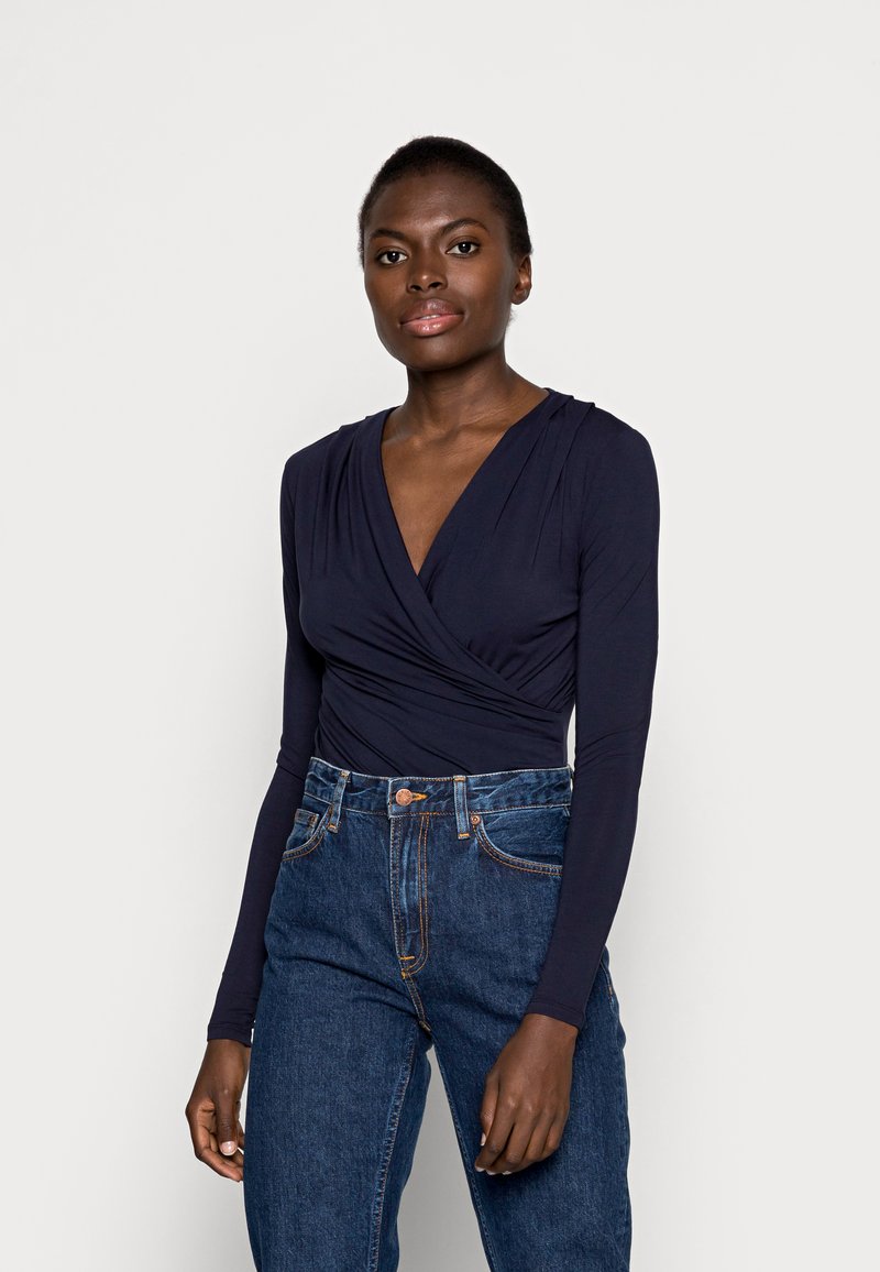 Mørkeblå langærmet indpakningstop med plisserede detaljer, parret med højtaljede blå jeans. Stoffet virker glat og blødt i teksturen.