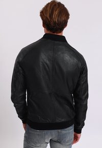 Man med ljusbrunt hår som bär en svart läderjacka och blå jeans, vänd bort från kameran mot en enkel bakgrund.