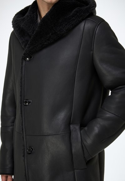 Schwarzer Ledermantel mit einer weichen schwarzen Pelzkapuze. Verfügt über Knopfverschlüsse und seitliche Taschen. Glatte Textur mit einer strukturierten Gestaltung.