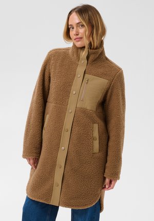 Femme portant un long manteau en polaire marron avec boutons-pression et poche poitrine zippée, debout devant un fond clair uni.