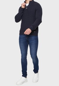 Maritimer Strickpullover mit strukturiertem Design, hohem Kragen und zwei Knöpfen. Kombiniert mit blauen Slim-Fit-Jeans und weißen Sneakers.