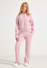 Ung kvinde står iført en lyserød velour zip-up hættejakke, matchende bukser og hvide sneakers foran en ensfarvet baggrund.