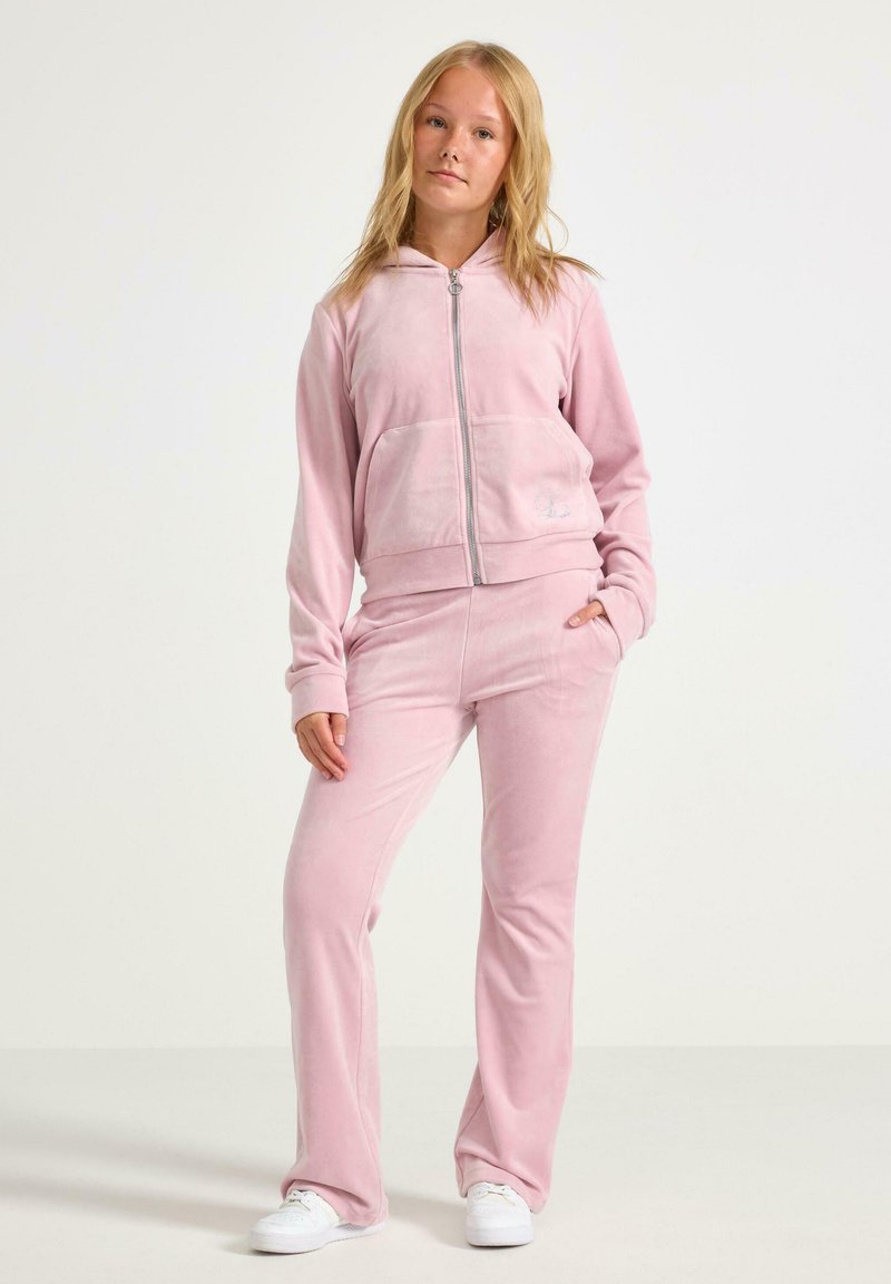Ung kvinde står iført en lyserød velour zip-up hættejakke, matchende bukser og hvide sneakers foran en ensfarvet baggrund.