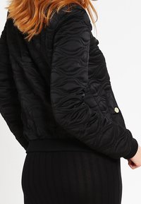 Czarna pikowana kurtka bomberka z teksturowanym wzorem, ściągaczami na rękawach i złotym guzikiem prasowanym. Gładki materiał o dopasowanym kształcie.