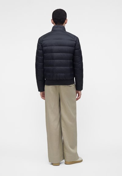Homme vu de dos portant une veste matelassée noire, un pantalon large beige et des chaussures claires sur un fond uni.