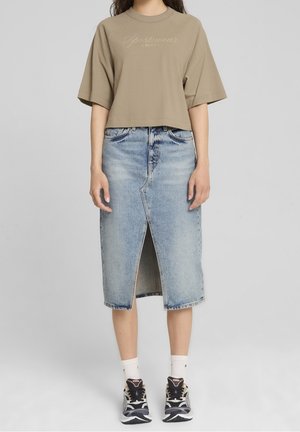 Person iført beige oversized t-shirt, lyseblåt denim midi-nederdel med frontslids, hvide sokker og sorte og hvide sportssko, stående på grå baggrund.